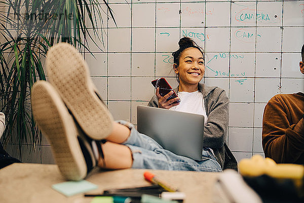 Smiling young businesswoman sitting with feet up on desk using wireless technologies by colleague against wall at office