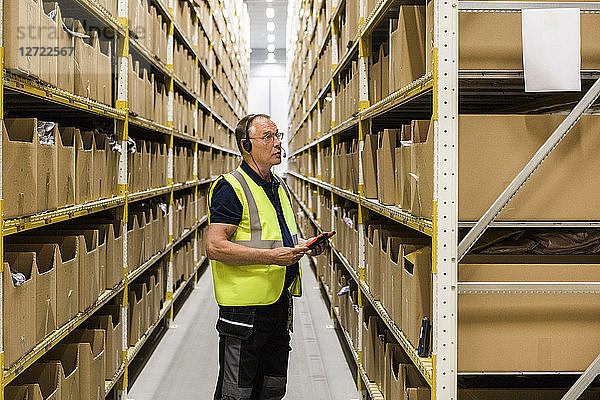 Side view of senior male worker with digital tablet looking at packages on rack while talking through headset at distrib