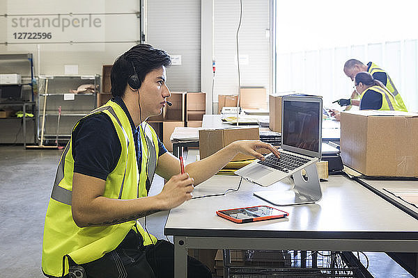Confident young male customer service representative looking away while sitting with laptop at desk in warehouse