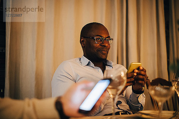 Smiling mature man using mobile phone while sitting with friend at dining table