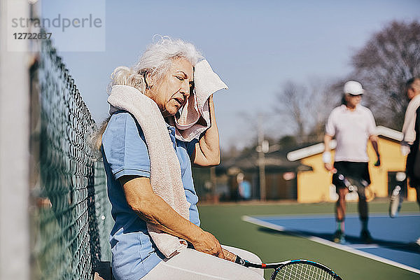 Exhausted senior woman wiping face with towel while sitting at tennis court