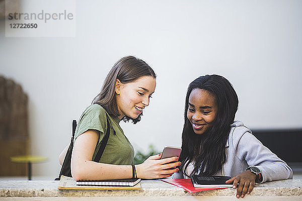 Smiling multi-ethnic female students sharing smart phone while standing in corridor at high school