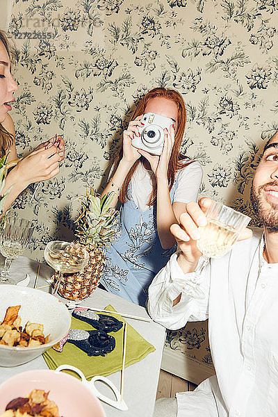 Young woman photographing while sitting with multi-ethnic friends at dining table during party in apartment