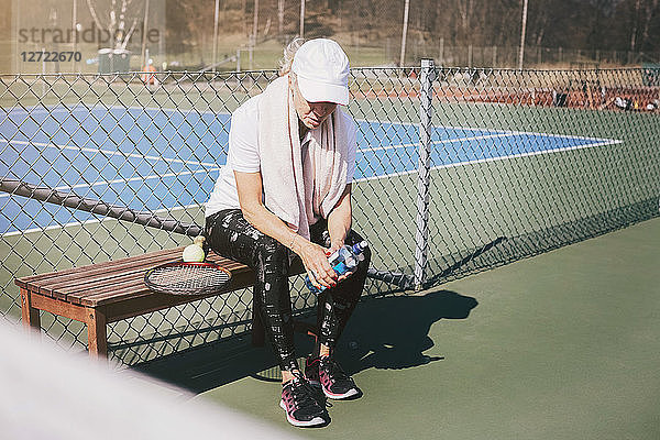 Full length of tired woman sitting on bench against net at tennis court