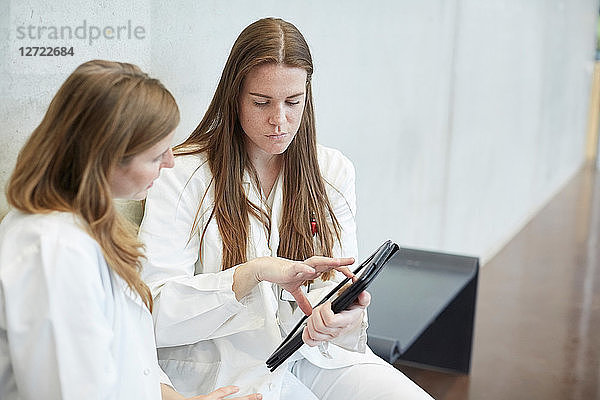 Confident female doctors discussing over digital tablet while sitting in corridor at hospital