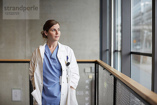 Thoughtful female doctor standing with hands in pockets while looking through window at hospital corridor