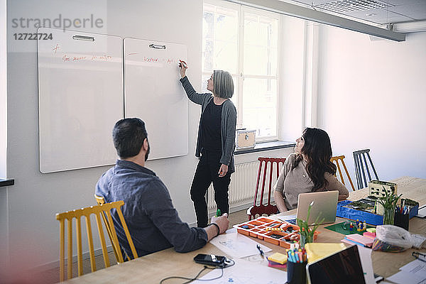 Female engineer writing on whiteboard while explaining colleagues sitting at table in office
