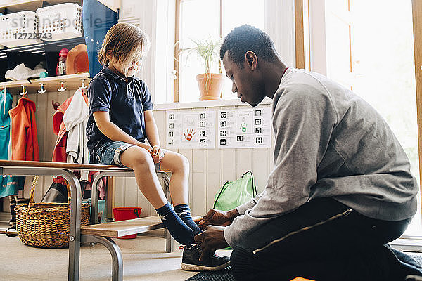 Side view of male teacher helping boy in wearing shoe at kindergarten