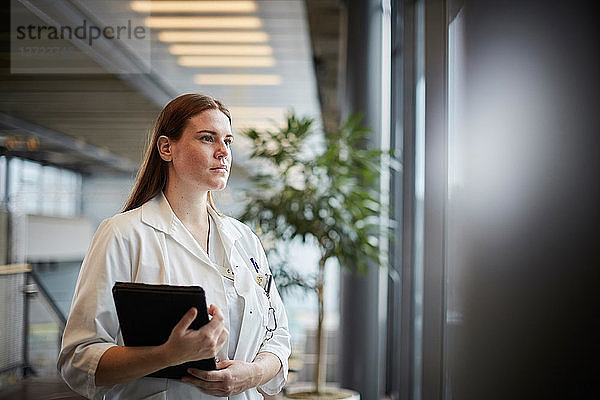 Thoughtful young female doctor with digital tablet looking through window at corridor in hospital