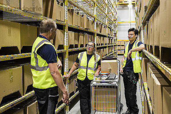 Multi-ethnic coworkers discussing while standing with cart on aisle amidst racks at distribution warehouse