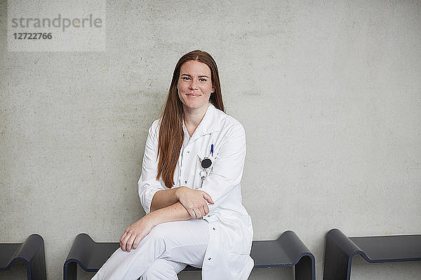 Portrait of smiling young female brunette doctor sitting against wall at hospital