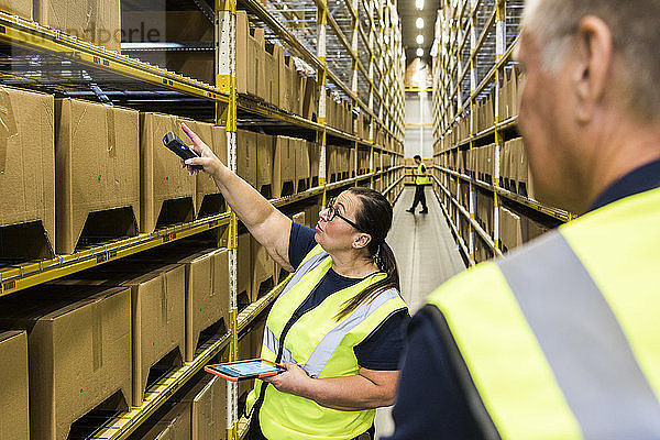 Female worker holding digital tablet while pointing at cardboard box on rack to coworker in distribution warehouse