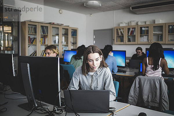 Confident female high school student using laptop at desk against teacher and friends sitting in computer lab