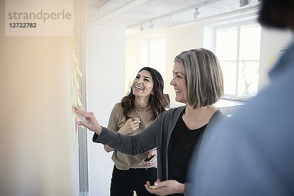 Mature female engineers smiling while reading adhesive notes stuck on glass in office