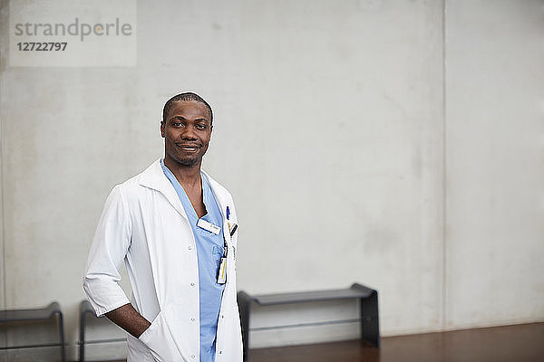 Portrait of confident male doctor standing with hands in pockets against wall at hospital