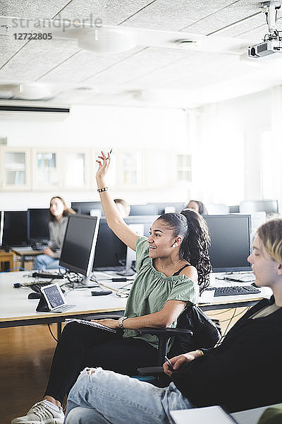 Smiling teenage girl sitting with hand raised by young friend in computer lab at high school