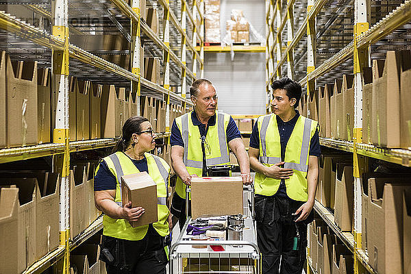 Multi-ethnic coworkers discussing while walking with cart on aisle amidst racks at distribution warehouse