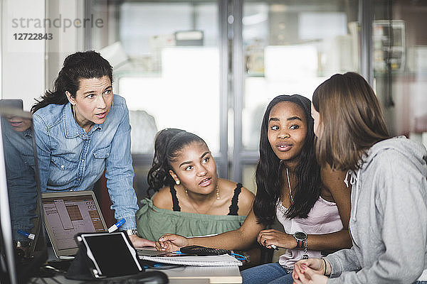 Mature teacher looking at confident female high school students discussing over laptop in computer lab