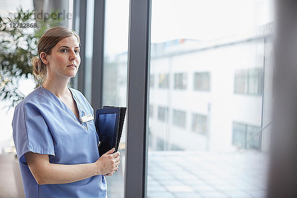 Thoughtful female nurse looking through window while standing in corridor at hospital