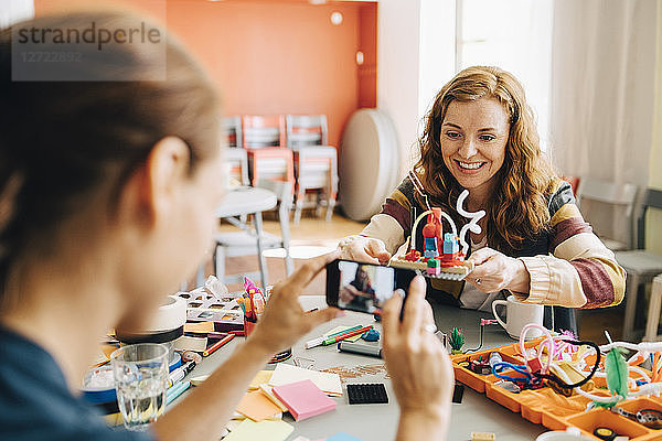 Businesswoman photographing smiling female colleague holding toy at table in creative office