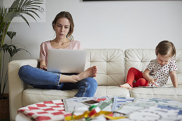Fashion designer looking at laptop while daughter using digital tablet on sofa at home