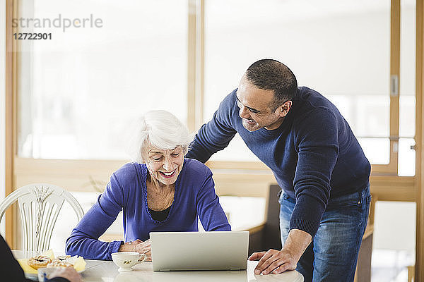 Cheerful mature man standing by mother looking at laptop on table in nursing home