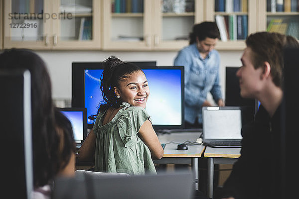 Smiling female high school student looking at friend sitting in computer lab