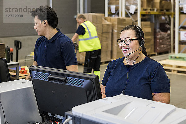 Smiling mature female customer service representative talking through headset while standing by coworker in distribution