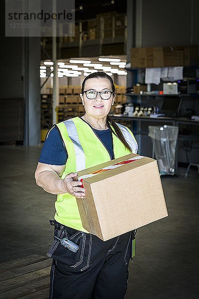 Portrait of confident mature female worker holding cardboard box while standing at distribution warehouse