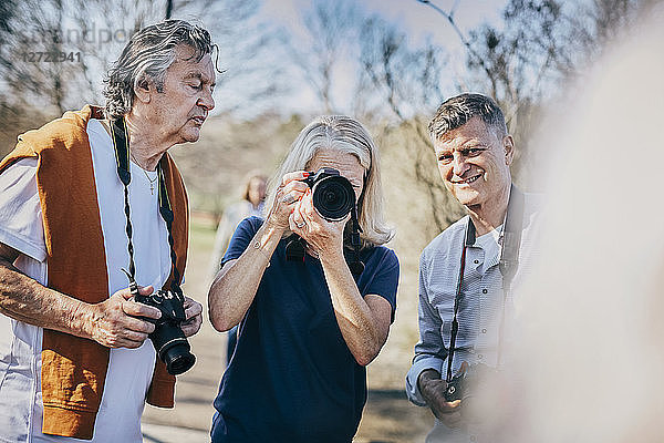 Senior woman using camera while standing with friends against bare trees at park