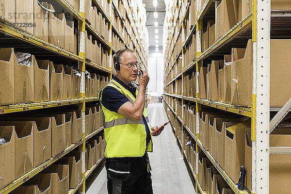 Senior male worker with digital tablet looking at packages on rack while talking through headset at distribution warehou