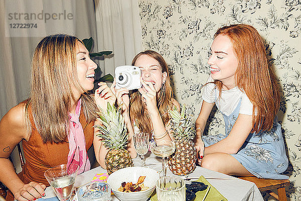 Cheerful young woman holding camera while sitting amidst female friends at home during dinner party