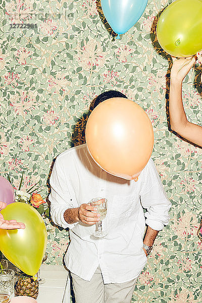 Obscured face of young man holding drink while standing by woman with balloons against wallpaper at home during dinner p