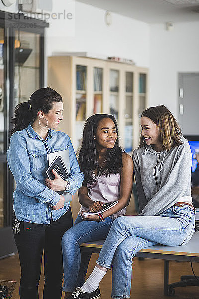 Smiling female teacher looking at high school students sitting on desk in computer lab