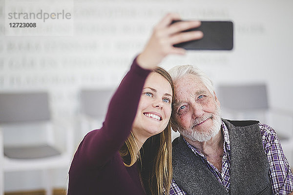 Smiling young woman taking selfie with grandfather while sitting in nursing home