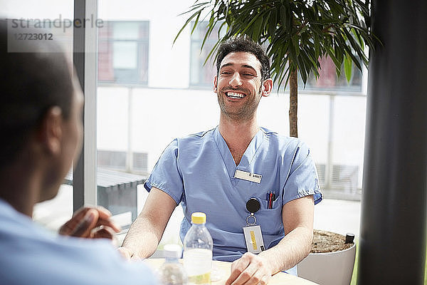 Smiling young male nurse talking with mid adult coworker against window at cafeteria in hospital