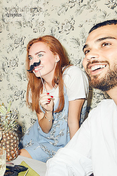 Smiling redhead woman holding mustache prop while sitting by male friend during party at home