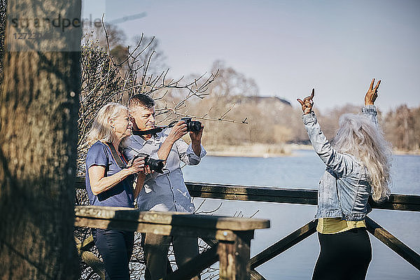 Senior man and woman photographing friend through camera while standing by lake