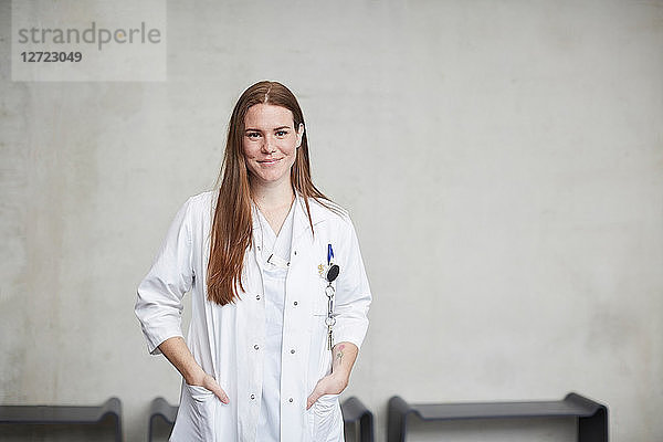 Portrait of smiling young female brunette doctor standing with hands in pockets at hospital