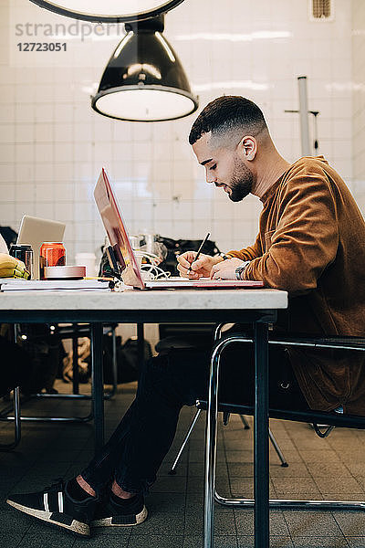 Full length side view of confident young male computer programmer writing while sitting with laptop at desk in small off