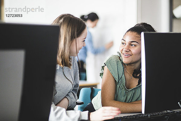Smiling multi-ethnic teenage girls discussing at desk in computer lab at high school