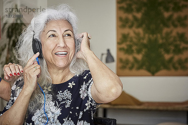 Smiling senior woman wearing headphones looking away while sitting with friend at home