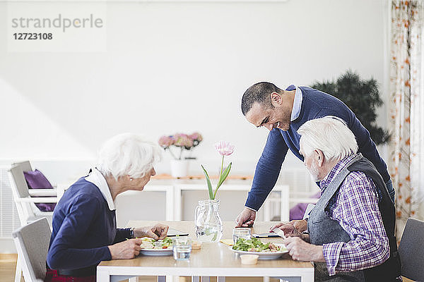 Son showing mobile phone to mother and father having lunch at table in nursing home