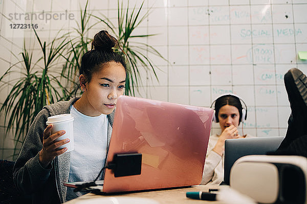 Confident young businesswoman holding coffee while using laptop by female colleague at creative office