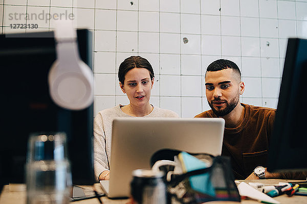 Confident young male and female colleagues looking at laptop against wall in office