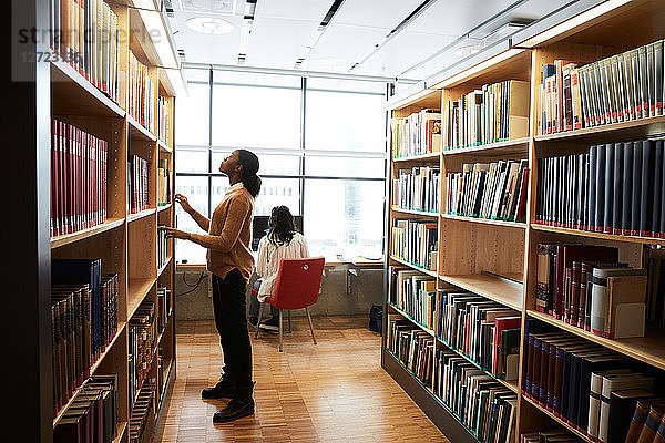 Full length of woman searching book on shelf while friend using computer in university library