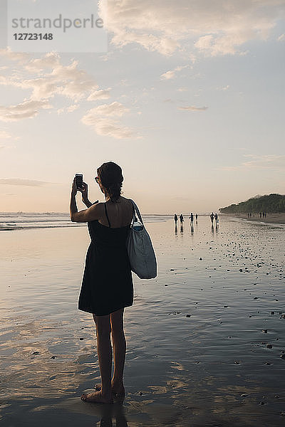 Full length rear view of mature woman photographing sunset using mobile phone at beach