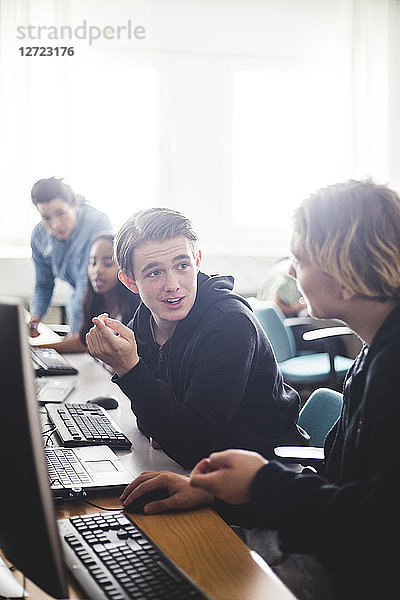 Male high school students discussing while sitting at desk using computers in classroom