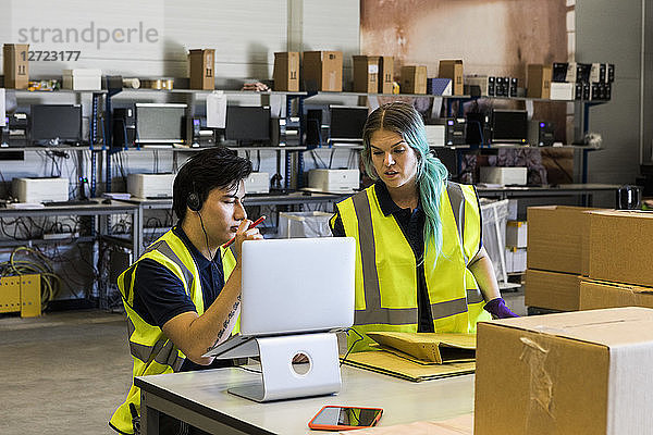 Confident young male customer service representative discussing with coworker over laptop while at desk in warehouse