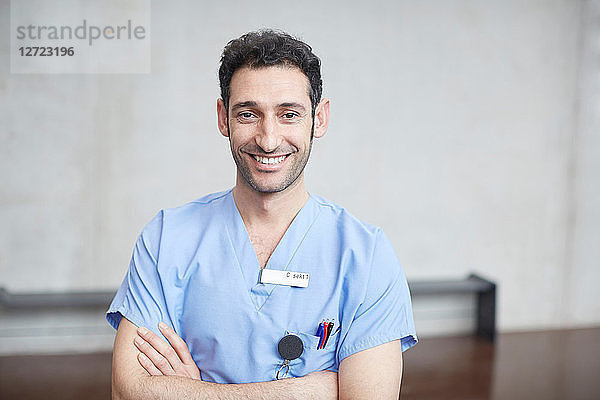 Portrait of smiling young male nurse in blue scrubs standing with arms crossed against wall at hospital